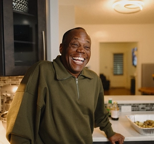 Smiling man in an olive green pullover standing in a kitchen, leaning on the counter.