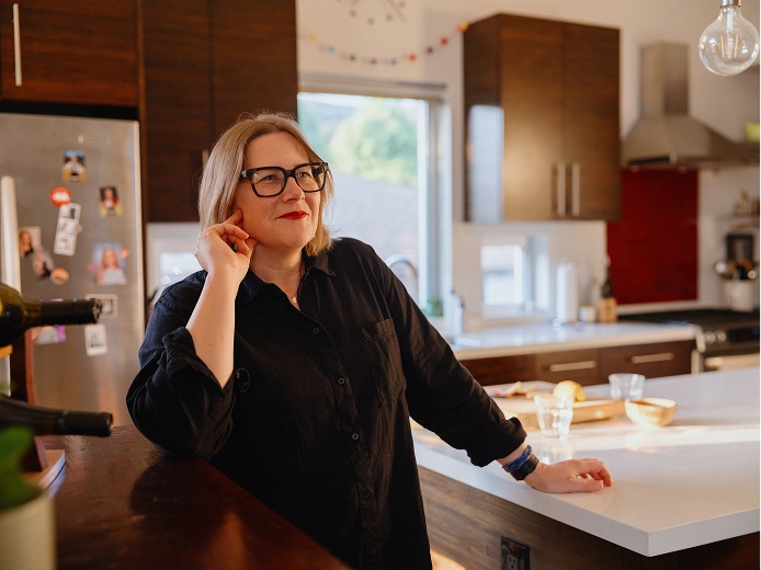 A woman wearing glasses and a black button-up shirt stands casually in a modern kitchen, leaning against a white countertop with one hand while resting the other near their face.