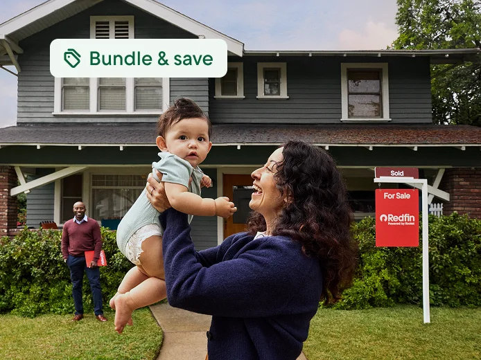 A woman lifting a baby in front of a green two-story house with a Redfin “Sold” sign in the yard while the realtor stands behind them. A tag says Bundle and save.